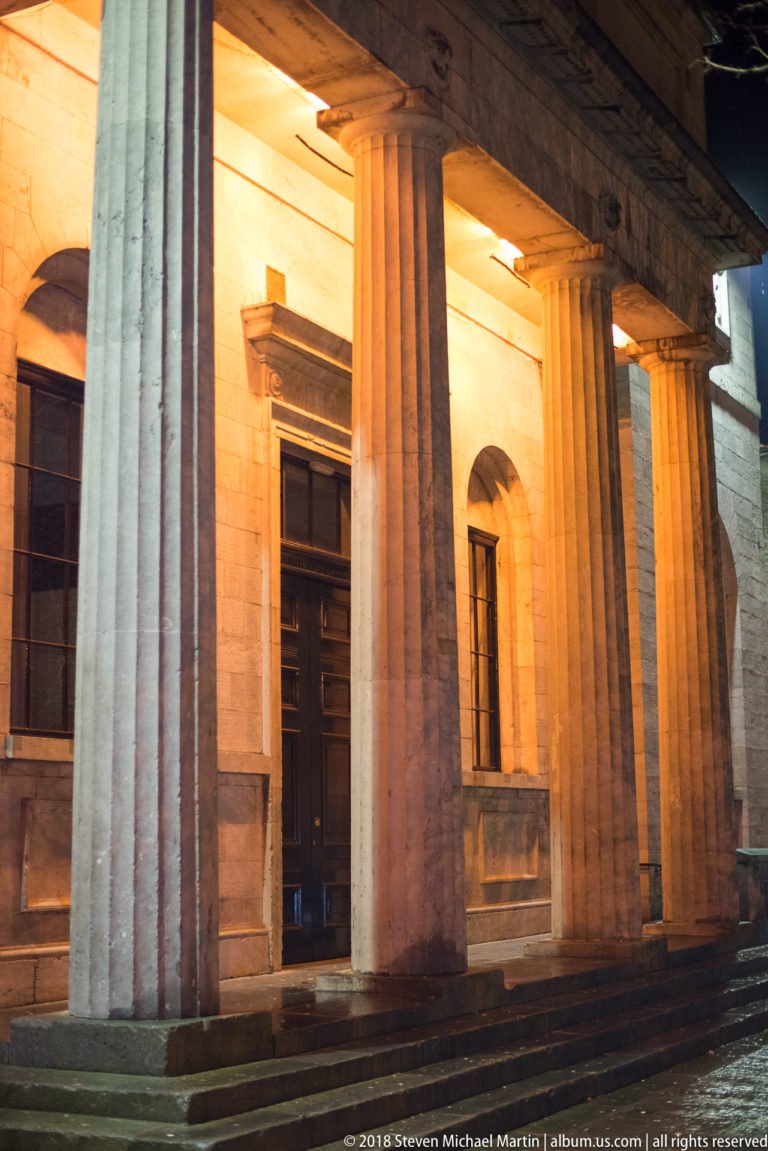 Floodlit civic building with tall fluted columns, recessed dark doors, and classical stone portico at night.