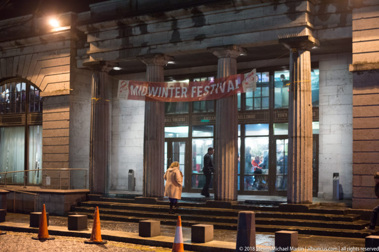 Town Hall Theatre entrance at night with Midwinter Festival banner, visitors, columns, glass doors, and traffic cones.