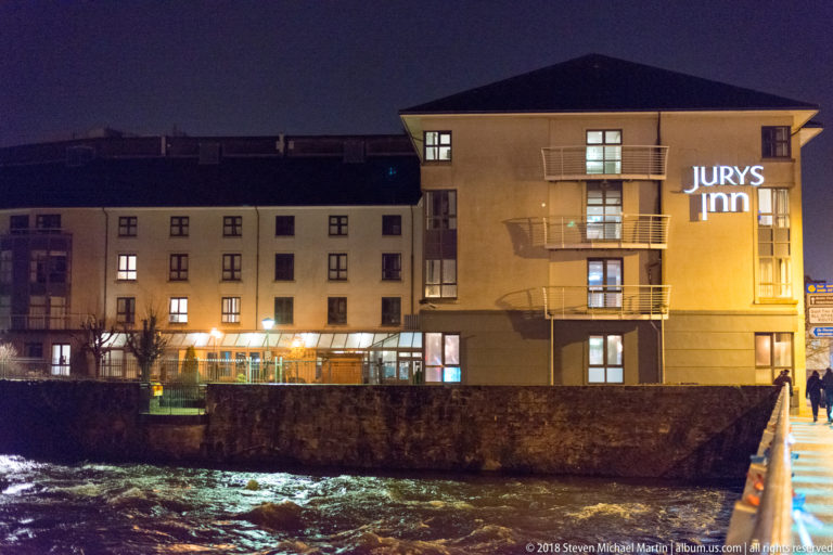 Jurys Inn and adjoining buildings behind a stone wall, with fast river water in the foreground at night.