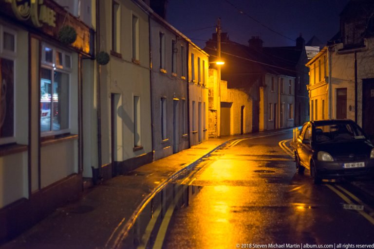 Narrow wet residential street curving between two-story houses, with parked cars and amber streetlights at night.