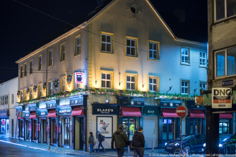 Blake's Bar on a lit Galway street corner with colorful awnings, pedestrians, neighboring storefronts, and festive lights.