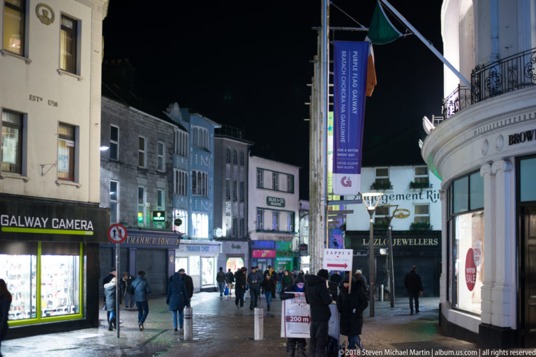 Pedestrians on a Galway shopping street at night, with camera shop, jewelers, signs, banners, and mixed historic façades.