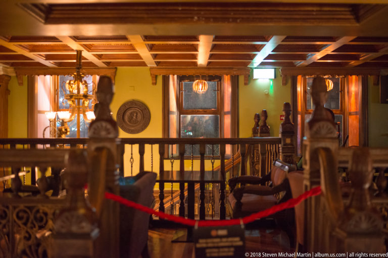 Warm traditional pub interior with dark carved wood, turned balusters, lamps, decorative figures, and a roped-off area.