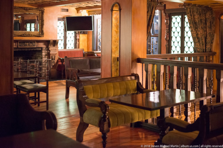 Traditional lounge with carved wooden tables, upholstered benches, patterned curtains, leaded windows, and warm lighting.