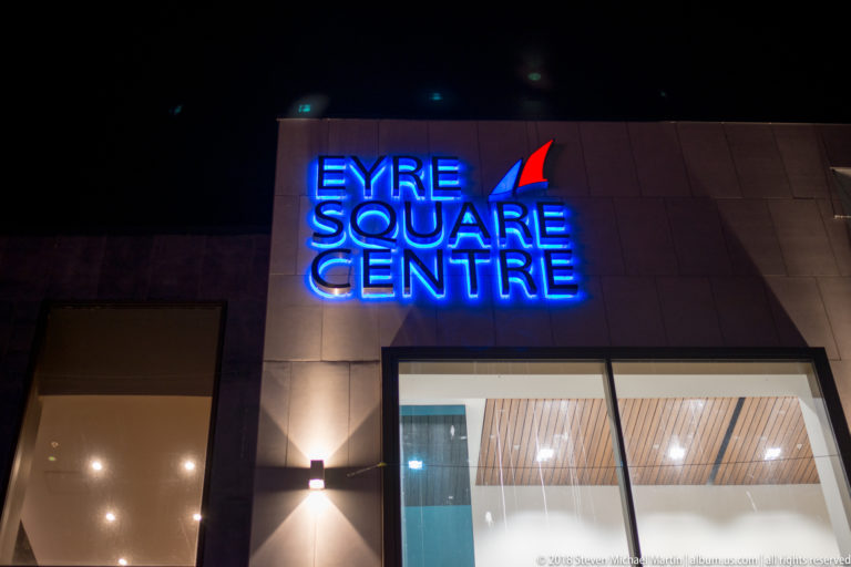 Blue neon EYRE SQUARE CENTRE sign above a modern glass entrance on a dark exterior façade.