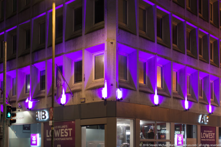 AIB bank building at night with purple uplighting on projecting concrete bays and illuminated street-level logos.