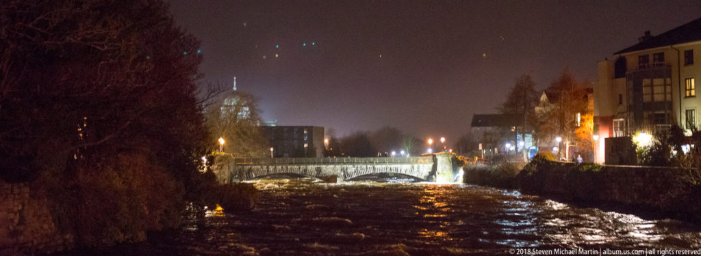Night view along the River Corrib toward a bridge and the illuminated dome of Galway Cathedral.