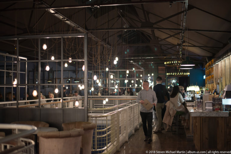 Industrial-style restaurant interior with white railings, hanging bulbs, timber bar, upholstered seating, and patrons gathered at the counter.