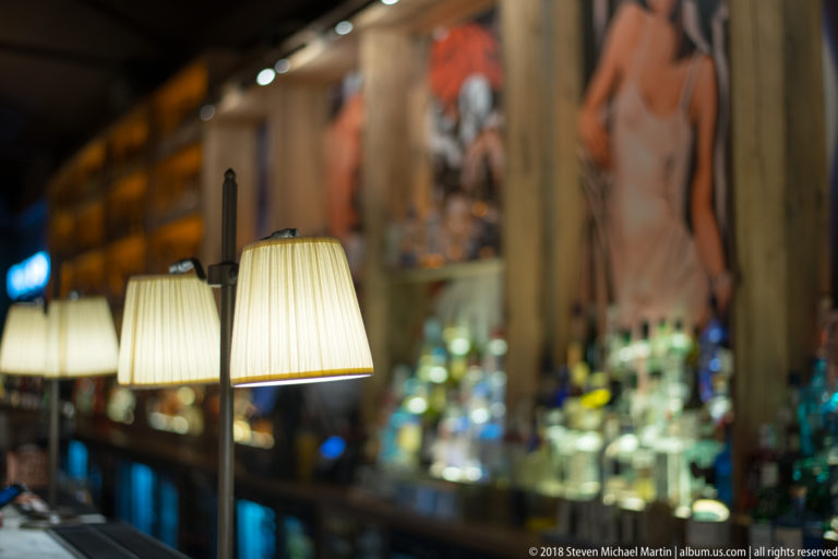 Pleated-shade lamps glow along a bar with blurred bottles and a large figurative mural in the background.