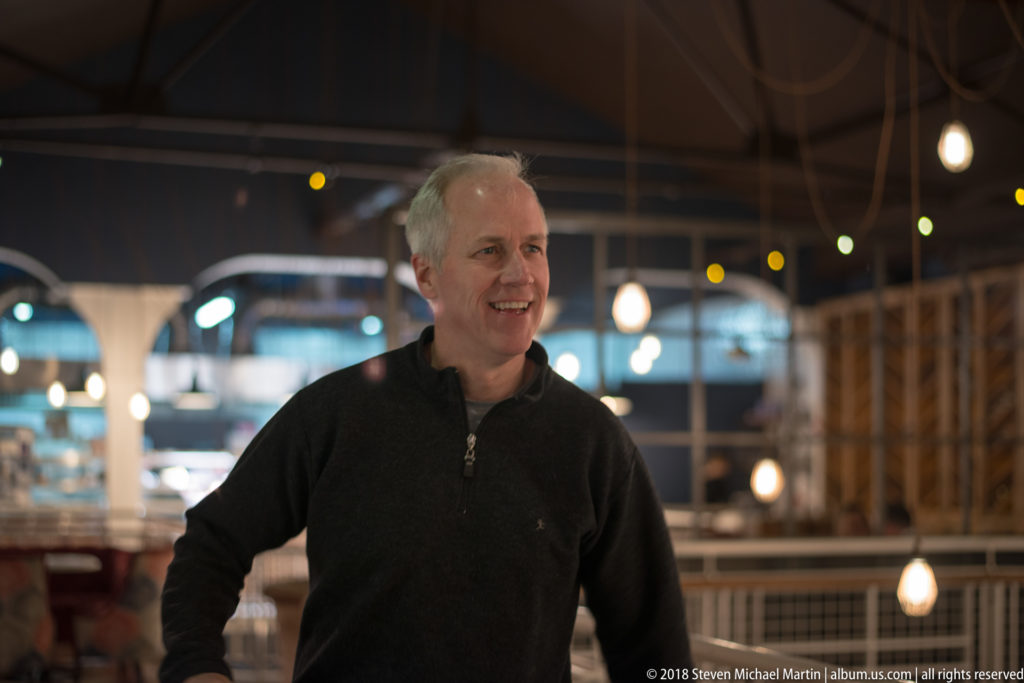 Smiling gray-haired man in a dark sweater standing inside a contemporary restaurant with pendant lights and railings behind him.