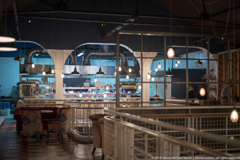 Open-kitchen restaurant interior with pale arches, pendant lamps, white railings, upholstered seating, and visible food-preparation area.