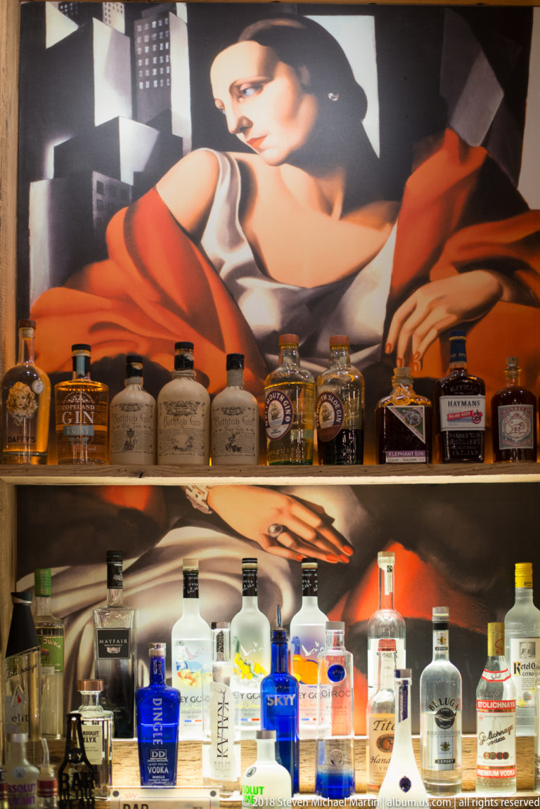Backlit spirit bottles on two shelves beneath a large mural of a woman dressed in red and white.