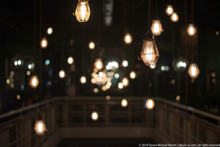 Warm filament bulbs in wire cages hang at varied heights above a dark interior railing with blurred lights beyond.