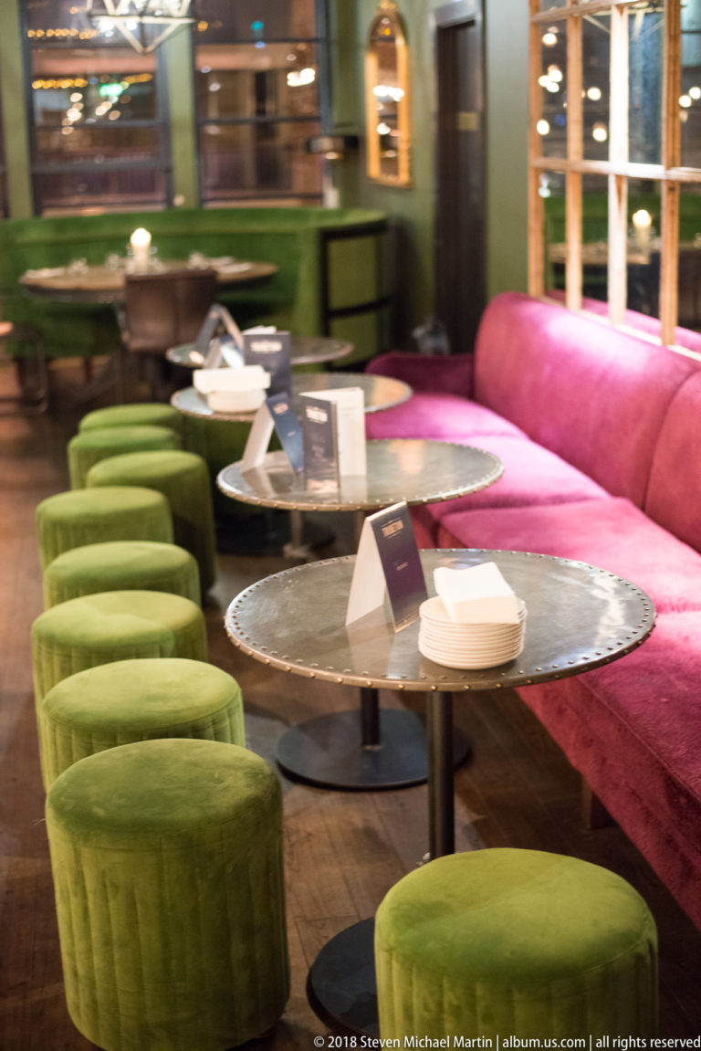 Green upholstered stools line round tables beside a long pink banquette in a colorful restaurant seating area.