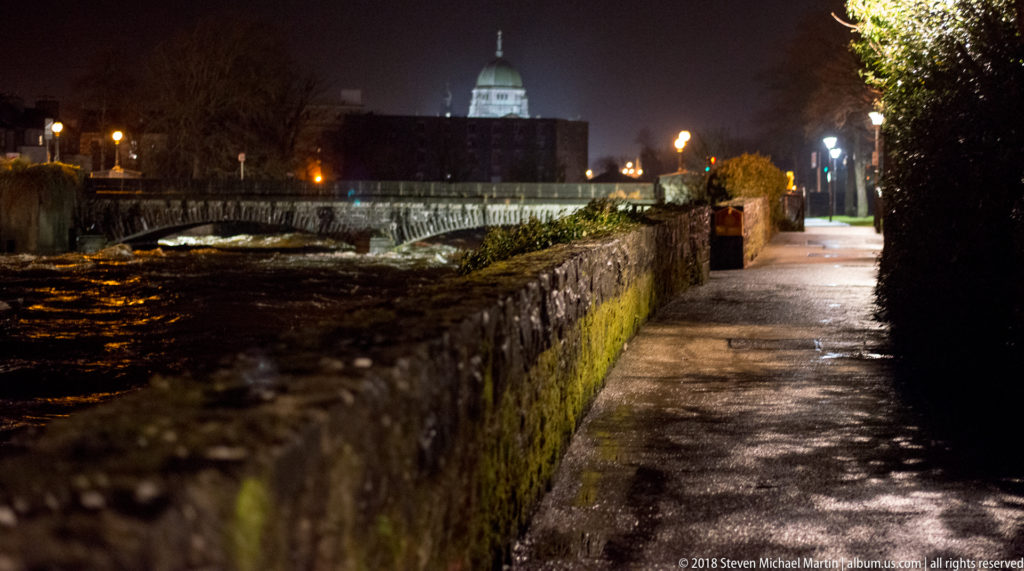 Wet riverside path beside a mossy stone wall, with bridge and Galway Cathedral dome visible ahead at night.