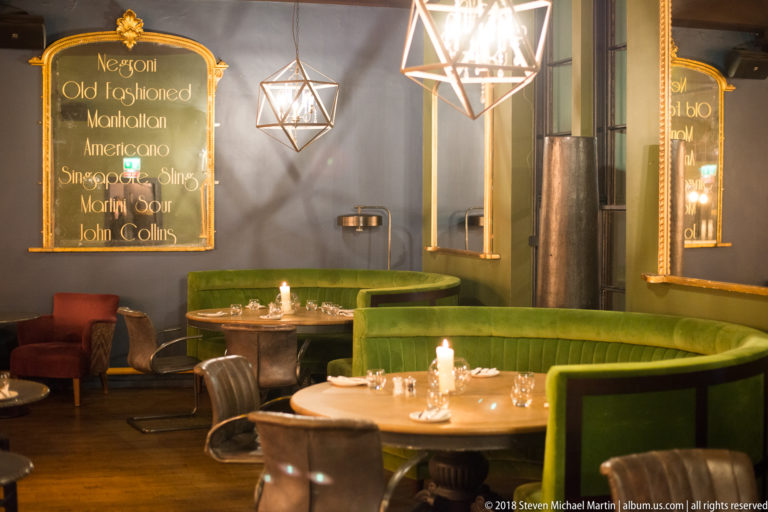 Green banquettes, round set tables, candles, and geometric pendant lights in an intimate restaurant lounge area.