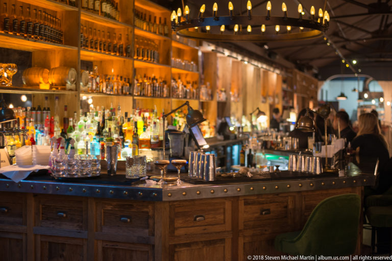 Large wooden cocktail bar crowded with bottles, glassware, mixing tools, warm lights, staff, and patrons.