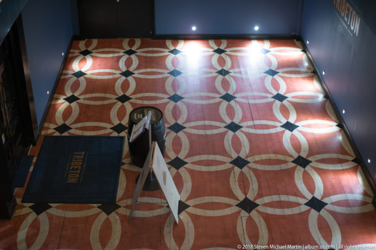 Geometric red, cream, and dark tiled floor with a TRIBETON mat, freestanding sign, and overhead spotlights.