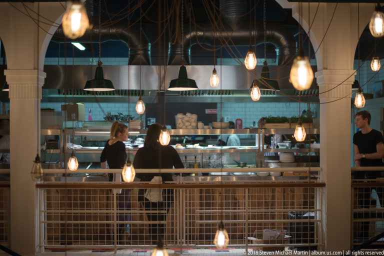 Restaurant staff working in an open kitchen behind pale columns, hanging lights, and a white metal railing.