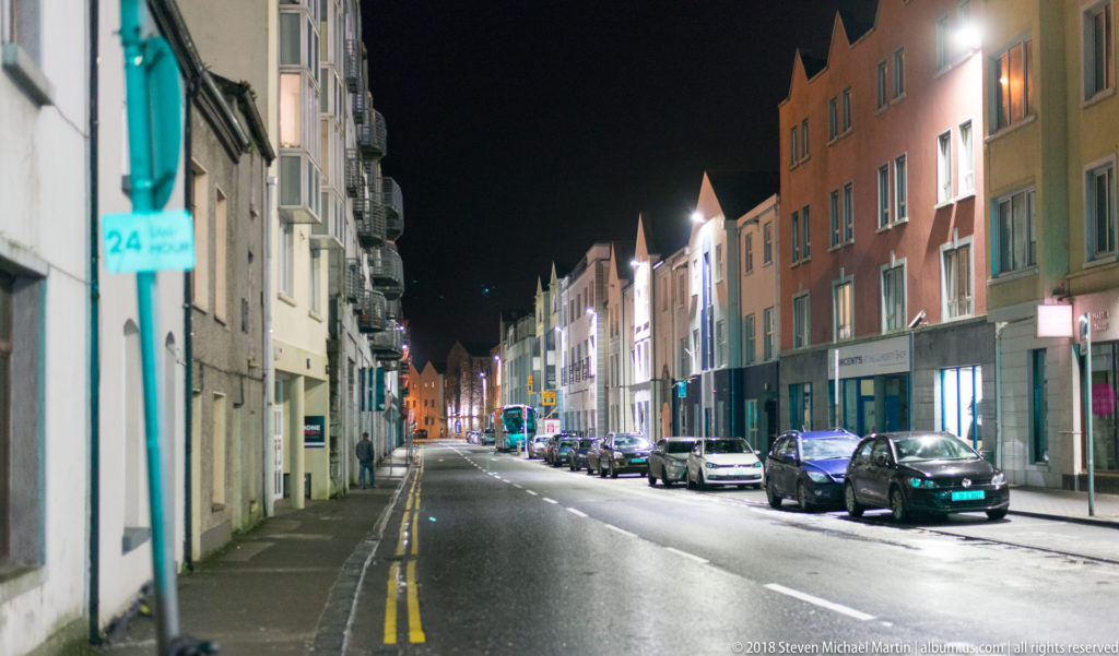 Quiet Galway street at night with parked cars, double yellow curb lines, and narrow multi-story buildings receding into the distance.