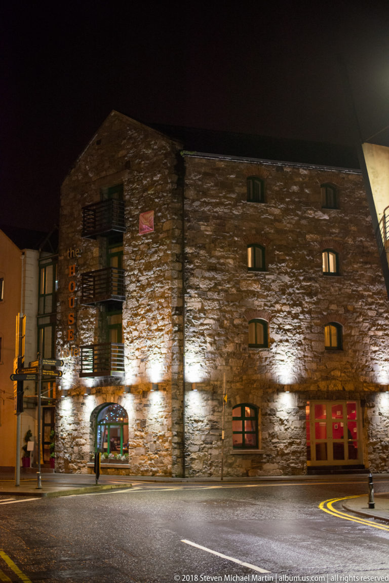 Large rough-stone building illuminated at night, with small upper windows and modern ground-floor commercial entrances.