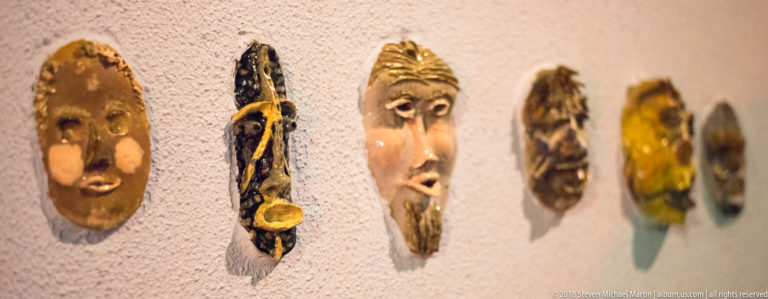 Row of small colorful sculptural masks mounted on a pale wall, with nearer faces in focus and others receding.