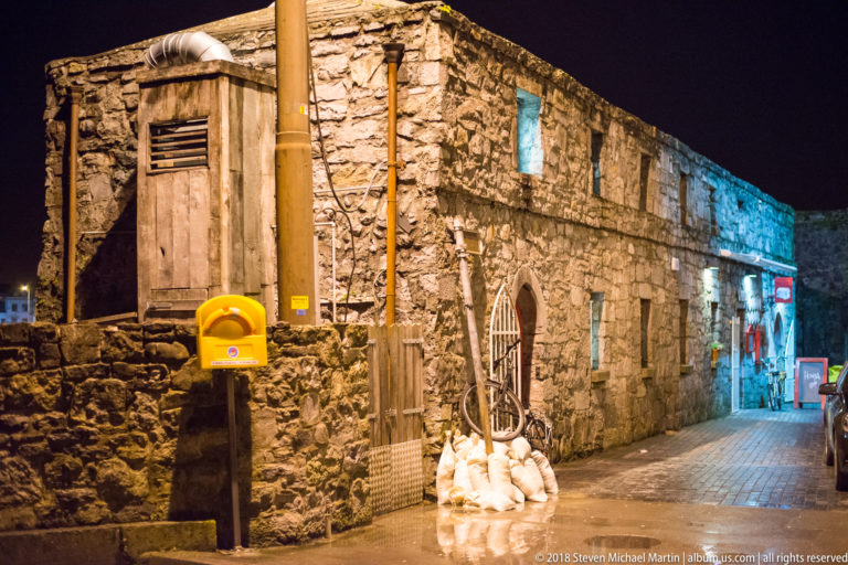 Rough stone building beside a narrow nighttime lane with pointed-arch gate, utility fixtures, and mixed warm and blue lighting.