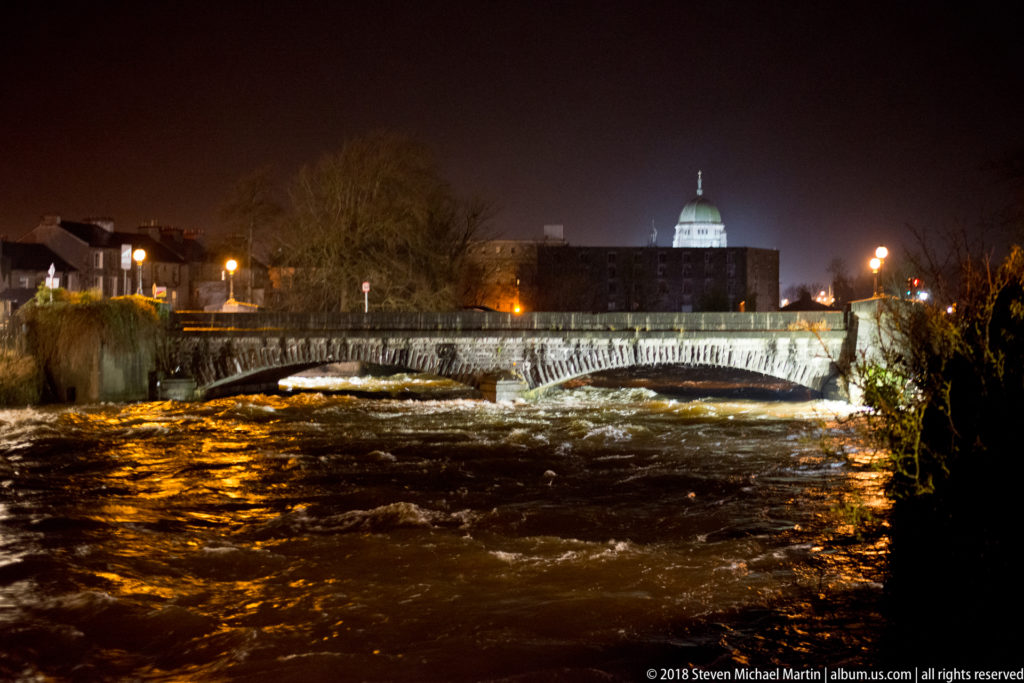 Fast-moving river water below a low bridge, with Galway Cathedral's illuminated dome in the background at night.