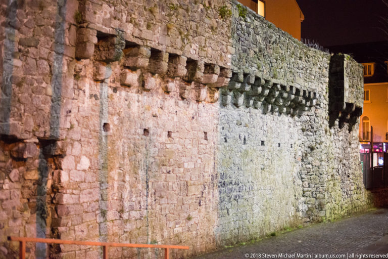 Massive rough-stone wall with a line of projecting corbels, seen obliquely beside a modern Galway street at night.