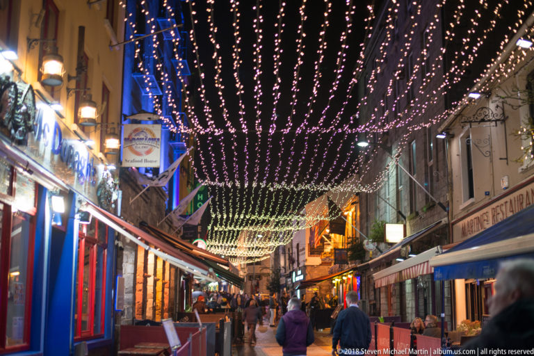 Pedestrians walk beneath a canopy of string lights on a narrow Galway street lined with pubs, shops, awnings, and signs.