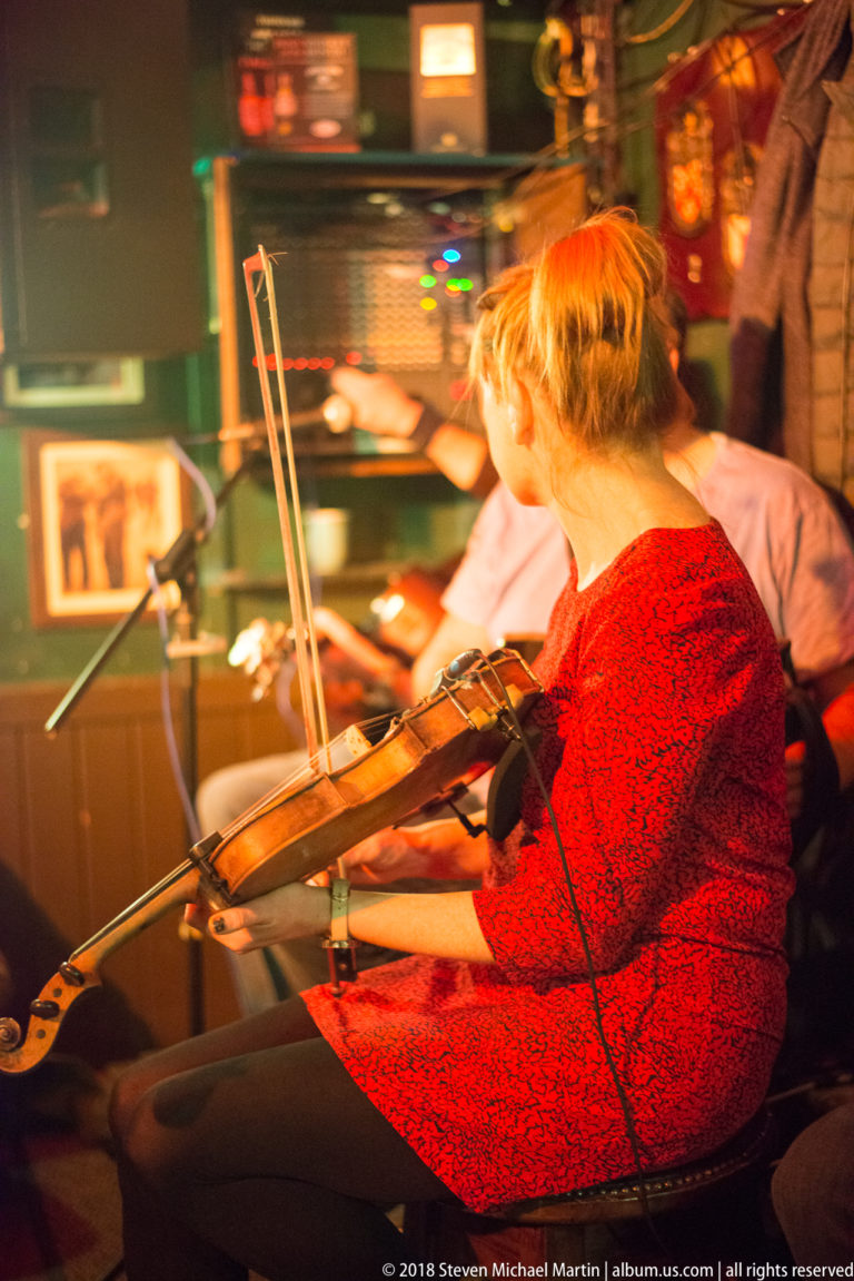 Woman in a red top playing fiddle in a warmly lit Galway pub, with microphones and another musician partly visible behind her.