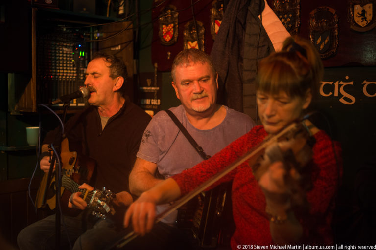 Three musicians performing closely together in a Galway pub, including a singer-guitarist and a fiddler in the foreground.