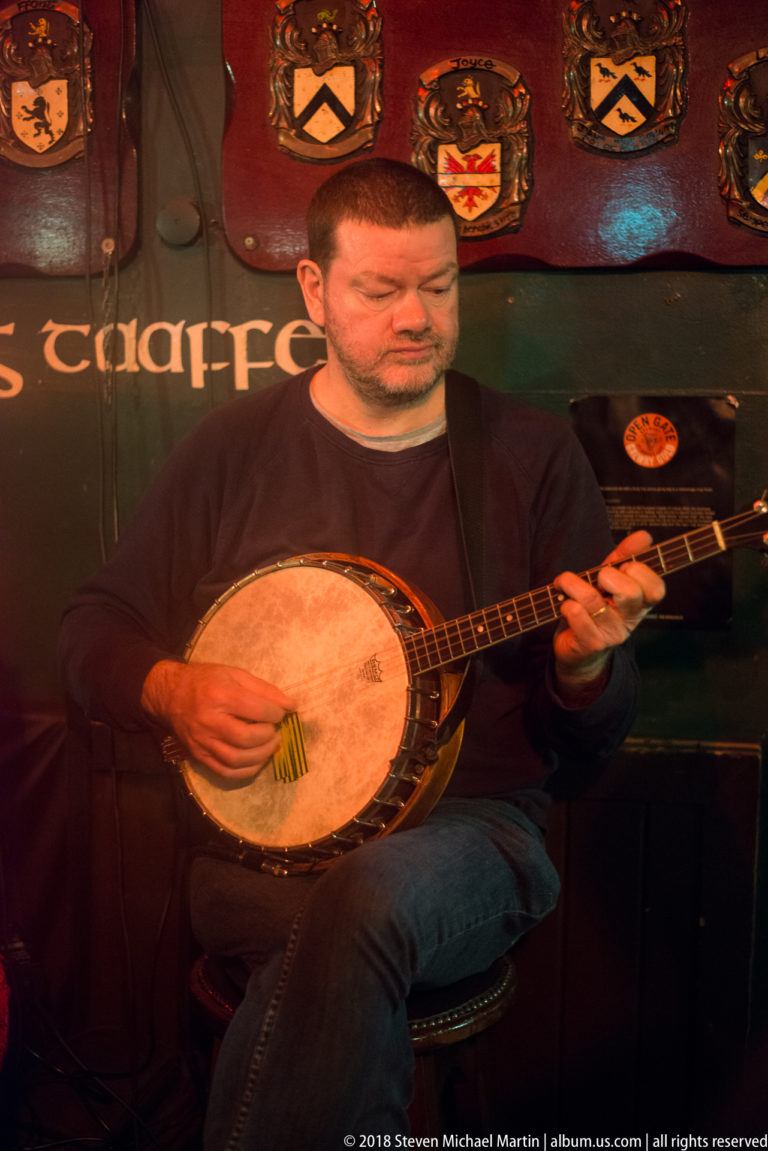 Seated male musician playing a four-string banjo beneath decorative shields in a warmly lit Galway pub.