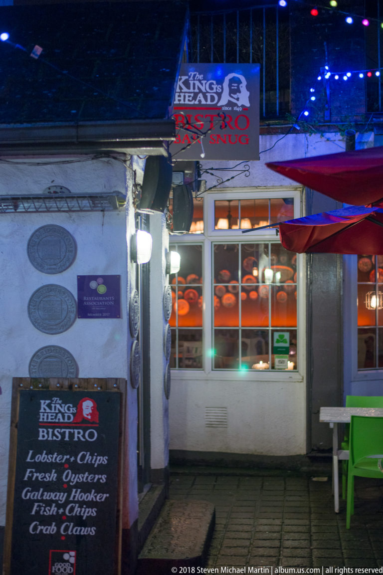 Night exterior of The Kings Head Bistro with illuminated windows, menu board, stonework, and colored lighting.