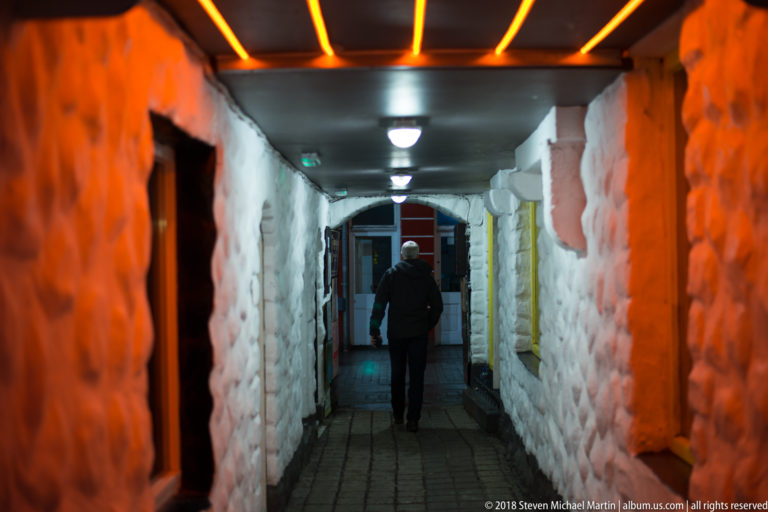Person walking through a narrow stone-walled corridor with evenly spaced ceiling lights and warm orange light on the foreground masonry.