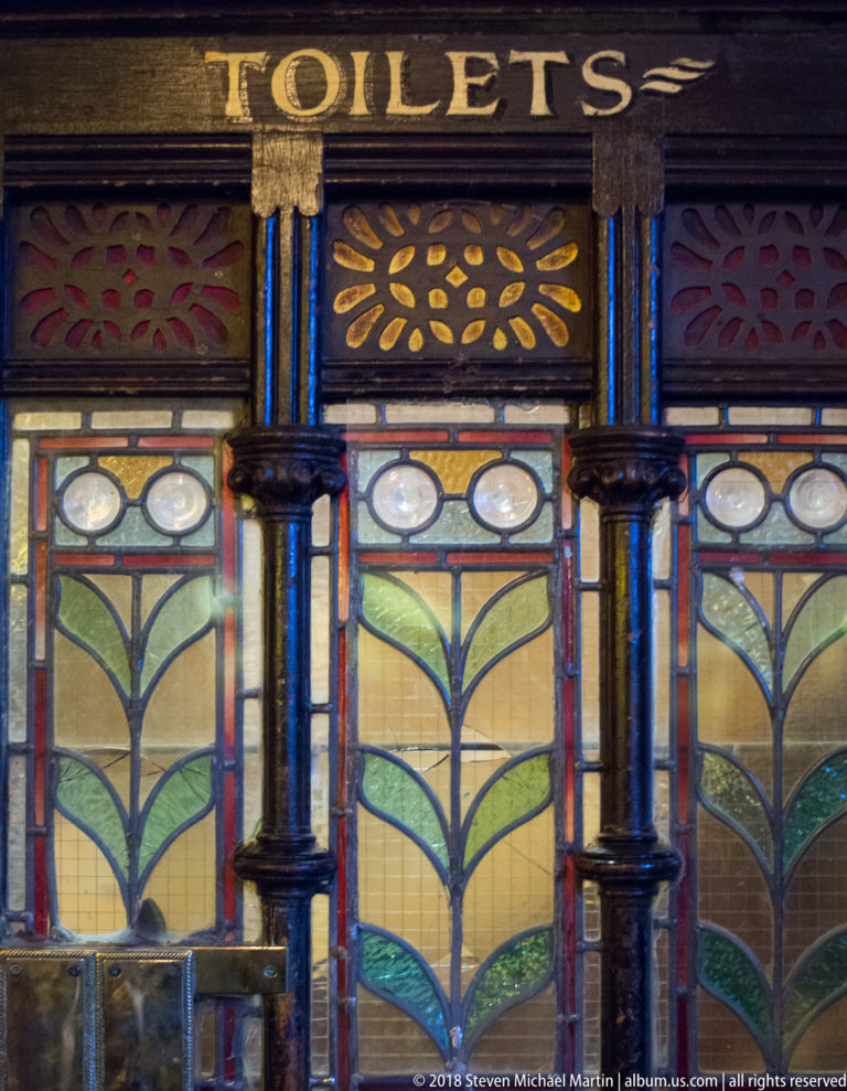 Decorative stained and textured glass panels beneath a carved TOILETS sign in a dark-wood pub interior.