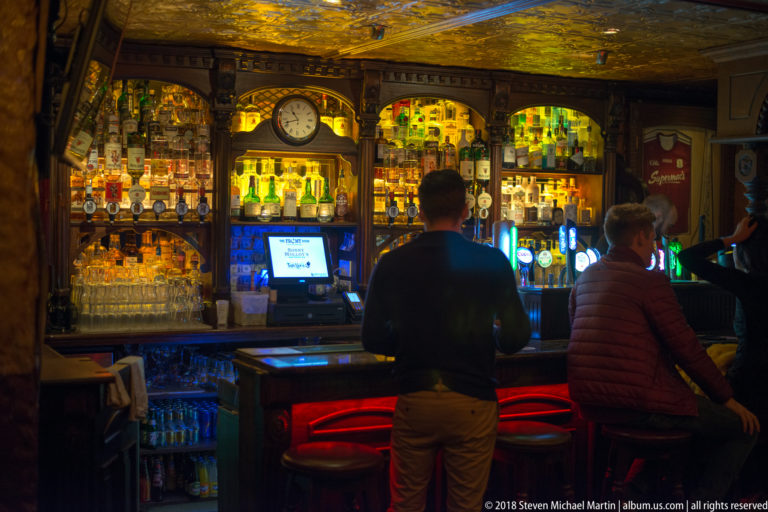 Two patrons seated at a traditional pub bar with illuminated spirit shelves, beer taps, dark wood arches, and a central clock.