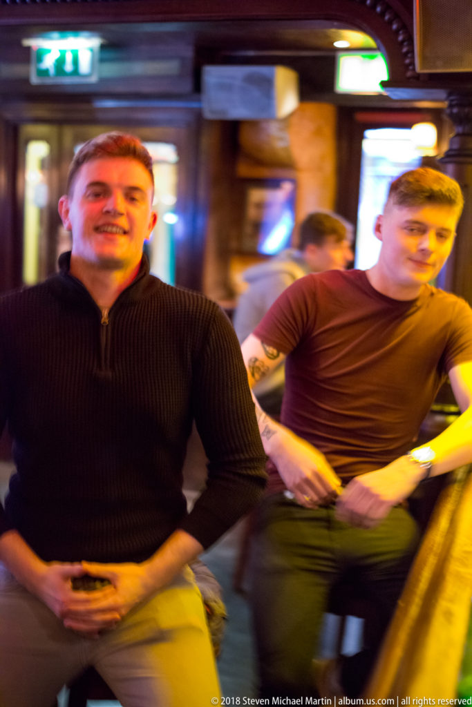Two men near a Galway pub bar with warm lighting, motion blur, and other patrons in the background.
