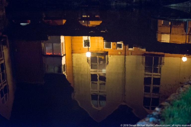 Upside-down reflection of a warmly lit building and lamp in dark water with gentle ripples.