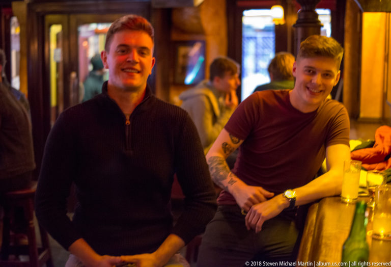 Two smiling men beside a Galway pub counter, with other patrons and warm interior lighting behind them.