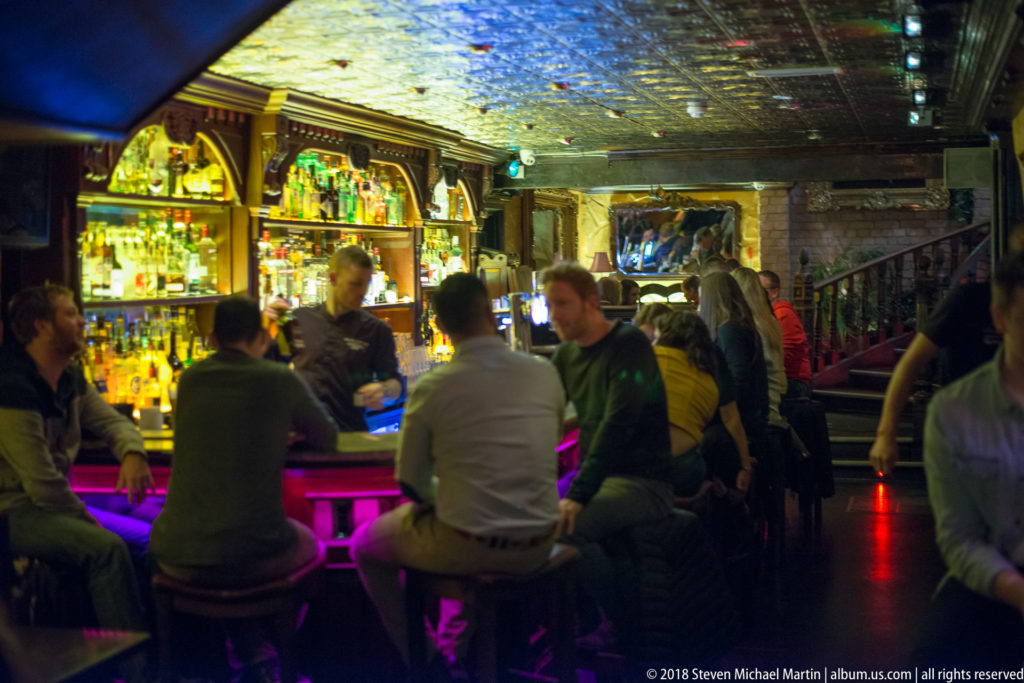 Crowded Galway pub interior with patrons seated at a brightly lit bar and bottles displayed behind the counter.