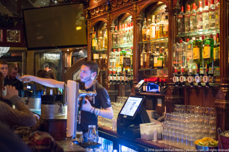 Bartender reaching across a pub counter beneath carved wooden shelving filled with liquor bottles, taps, and glassware.