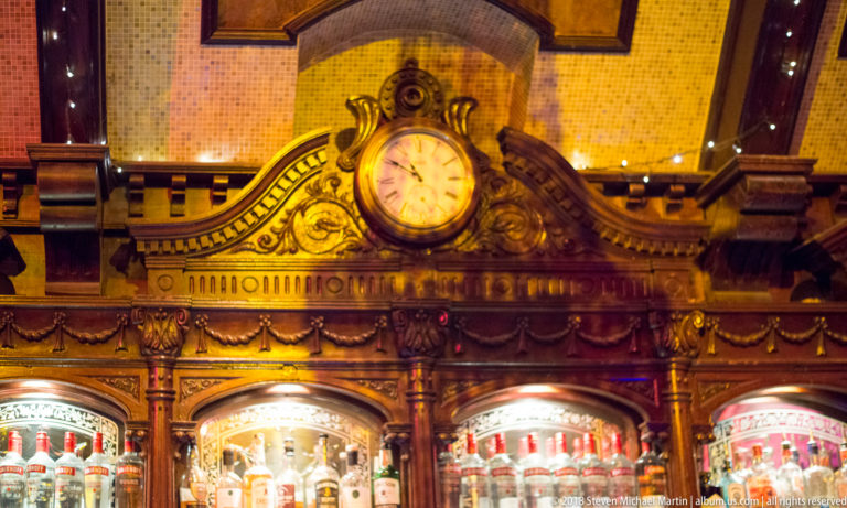 Round clock set into an ornate carved wooden bar surround above illuminated shelves of bottles.