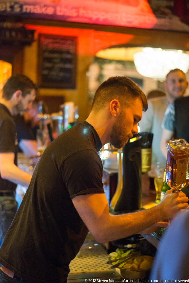 Bartender leaning over a busy pub counter with drink equipment and blurred patrons in the background.