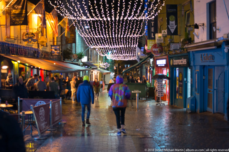 People walking along a wet Galway pedestrian street beneath dense strings of white decorative lights.