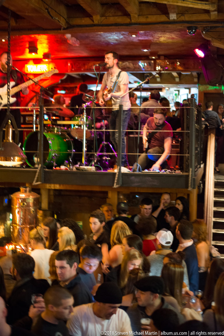Live band on an elevated pub stage above a dense crowd under warm red lighting.