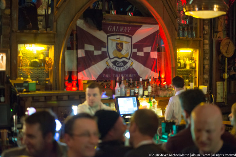 Maroon-and-white Galway GAA flag reading “The Tribesmen” hanging behind a crowded pub bar.