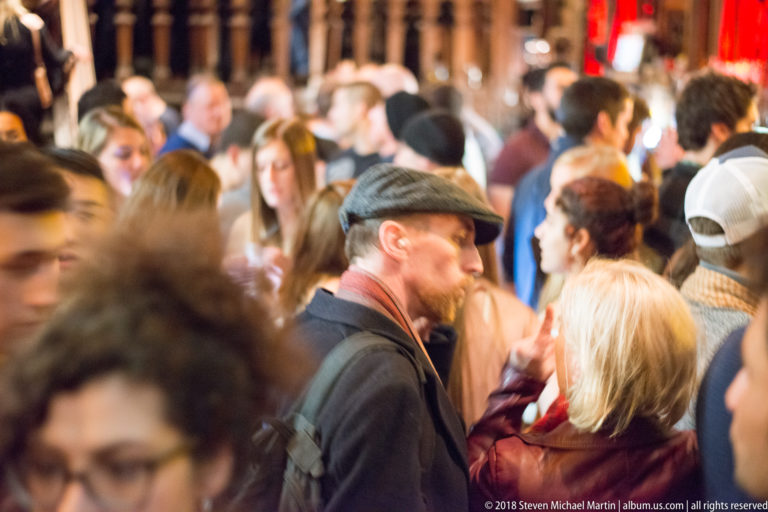 Dense Galway pub crowd with a man in a flat cap near the center and patrons surrounding him.