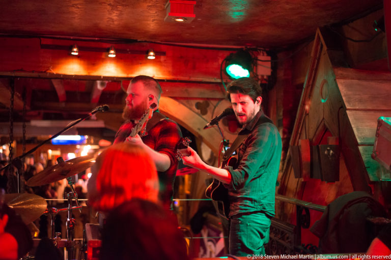Two guitarists performing close to the audience under red and green lights in a Galway pub.