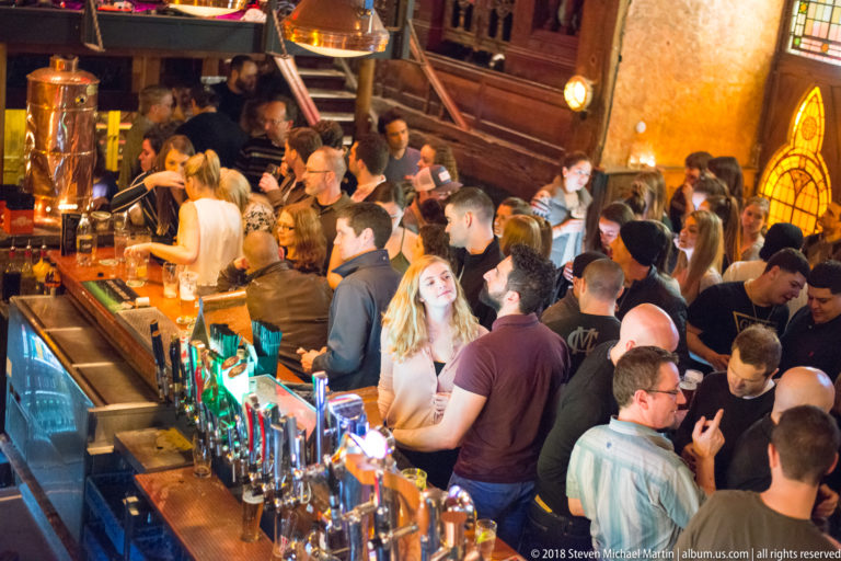 Elevated view over a busy Galway pub bar with staff, customers, beer taps, and tightly packed standing patrons.